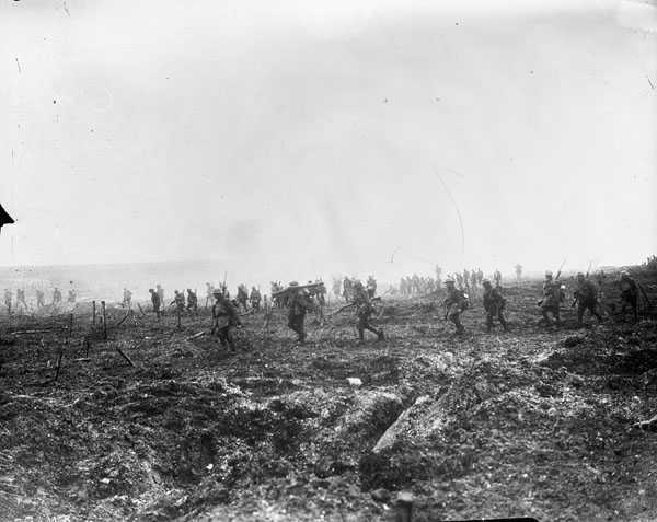 Black and white photograph. A large number of men walk, spaced out, through a misty day on very muddy and broken ground. Several carry large guns on their shoulders.